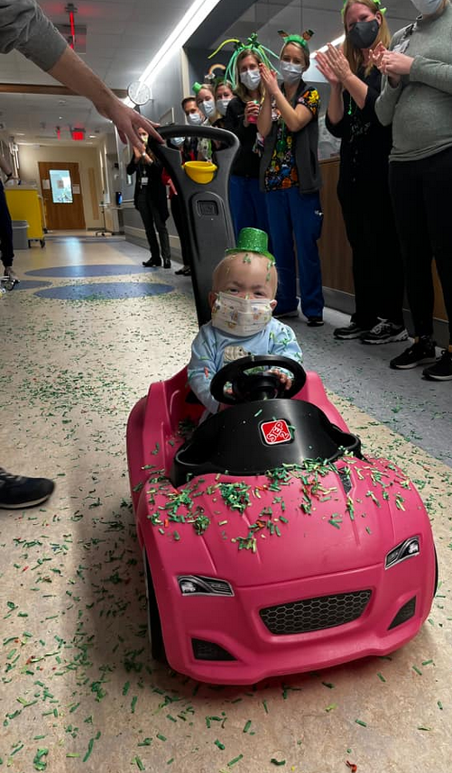 Small boy driving a toy car down a hospital hallway as the staff throws confetti and celebrates his recovery