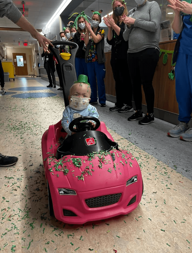 Small boy driving a toy car down a hospital hallway while staff celebrates and throws confetti