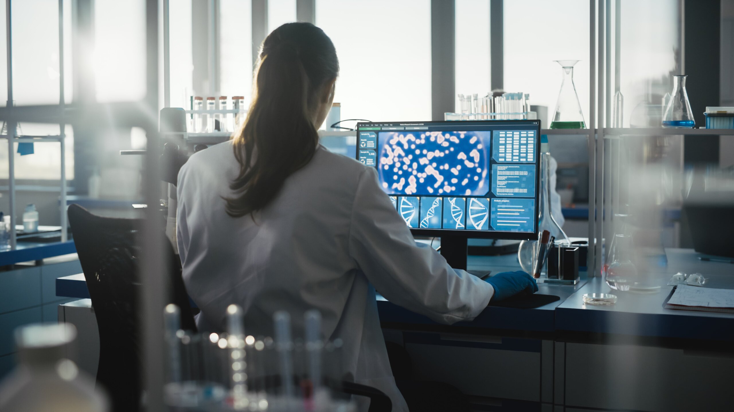 Woman in a lab coat sitting at a computer looking at DNA strands and items under a microscope